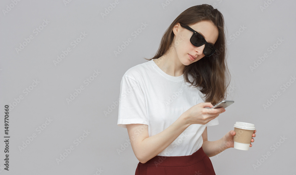 Studio portrait of hipster female isolated on gray background holding cardboard coffee cup from takeaway with head bent towards smartphone display watching media content with attention and interest.