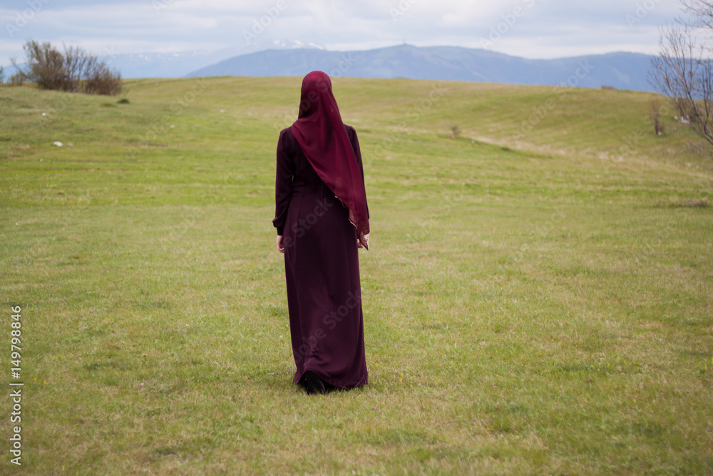 Young and beautiful girl covered, Muslim, in burgundy dress, walk through the meadows surrounding hills, enjoying the natural resources, relaxation Young and beautiful girl covered, Muslim, in burgundy dress, walk through the meadows surrounding hills, enjoying the natural resources, relaxation