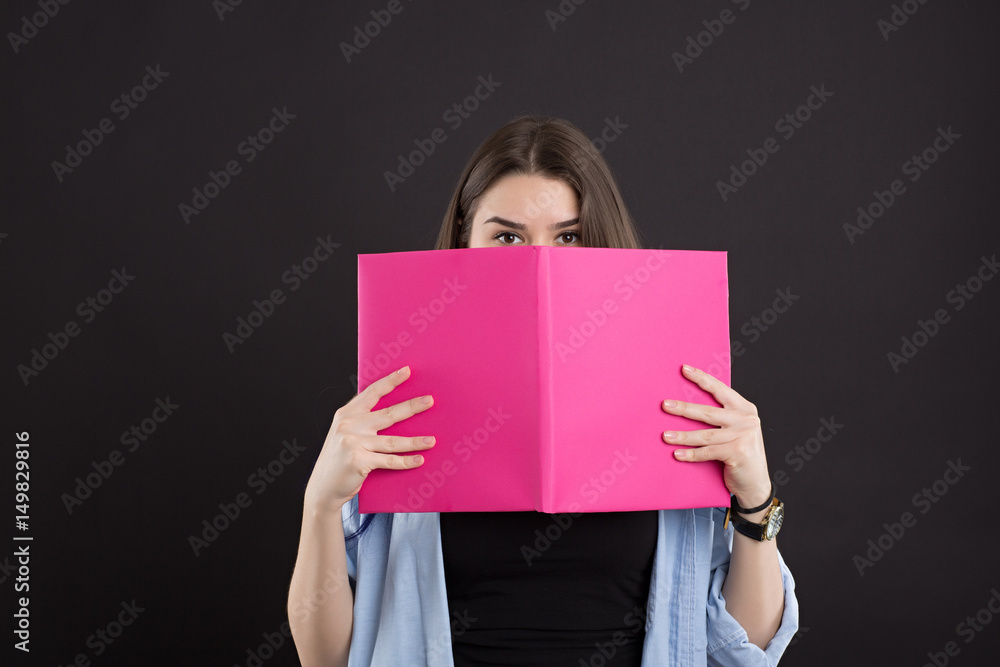 beautiful caucasian female student in denim shirt with long hair, over black background, covered her face with a book beautiful caucasian female student in denim shirt with long hair, over black background, covered her face with a book