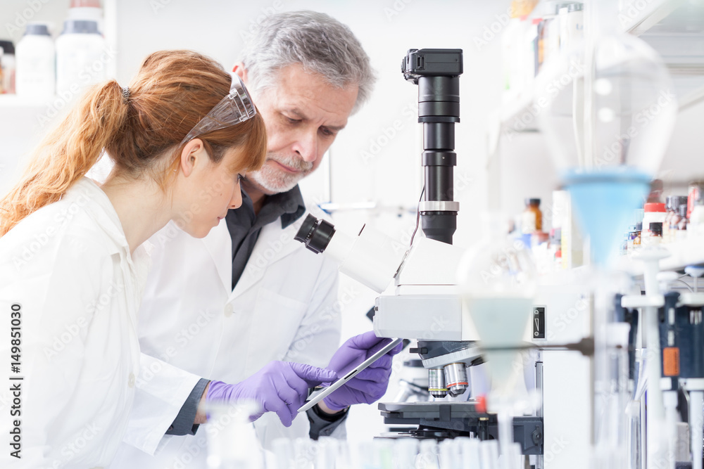 Health care researchers working in life scientific laboratory. Female research scientist and senior male supervisor looking focused at tablet computer screen evaluating and analyzing microscope image. Health care researchers working in life scientific laboratory. Female research scientist and senior male supervisor looking focused at tablet computer screen evaluating and analyzing microscope image.