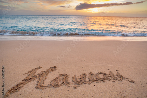 A Word «Hawaii» written on a sand of a tropical beach. Beautiful Sunset above the ocean water