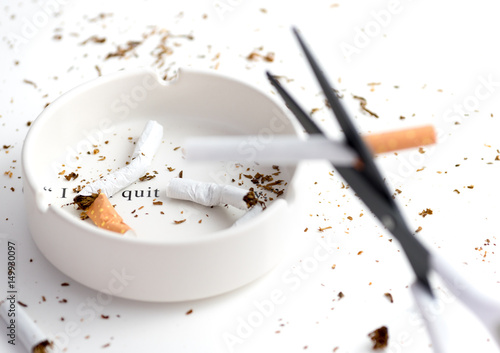 white ashtray with destroyed cigarette that emphasize words "i quit" from complete quote "i will quit tomorrow", blurred black blade scissors cutting cigarette as forground
