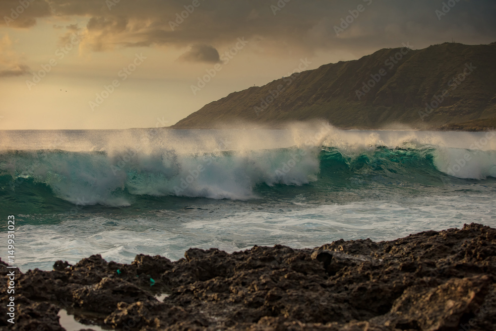 Beautiful green rough ocean wave incoming. Sunset sea with shorebreak water surface. Rocky coastline with mountain landscape on background