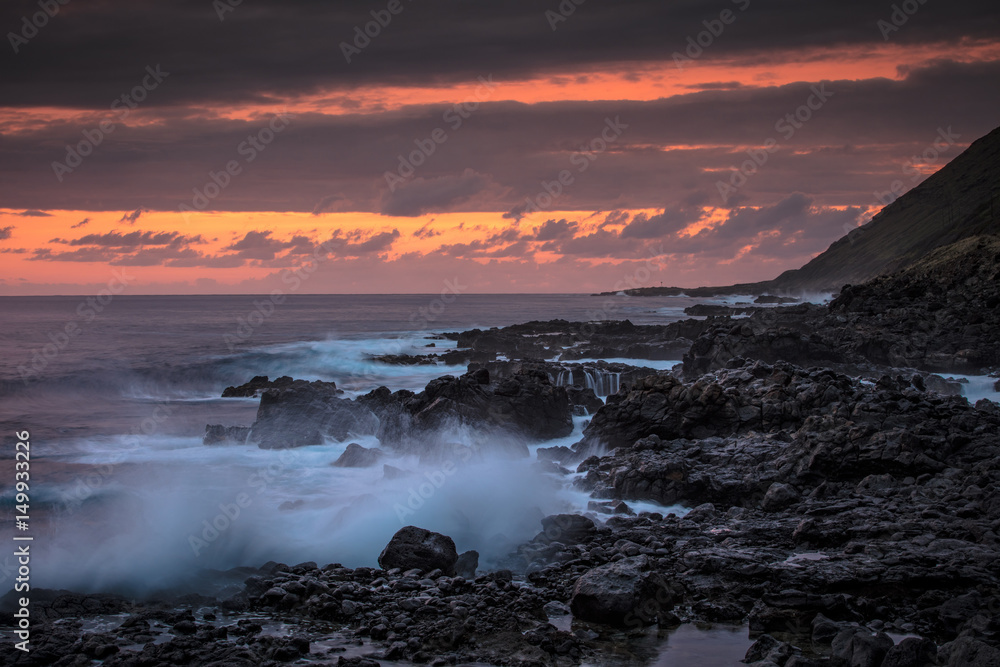 Beautiful sunset in ocean view with pinky orange sky on background. Rocky coastline with splashing against black lava rocks waves