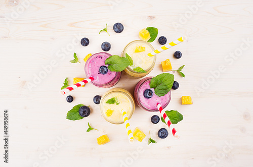 Fototapeta Naklejka Na Ścianę i Meble -  Freshly blended yellow and violet  fruit smoothie in glass jars with straw, mint leaves, mango slices, berry, top view. Soft white wooden board background.