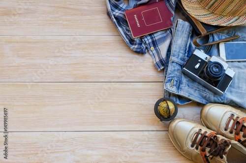 Top view of traveler´s accessories: leather bag, blue jeans, hat, check shirt, gold sneakers, smart phone, vintage camera and passport on wooden background with space for text. Travel outfit concept.
