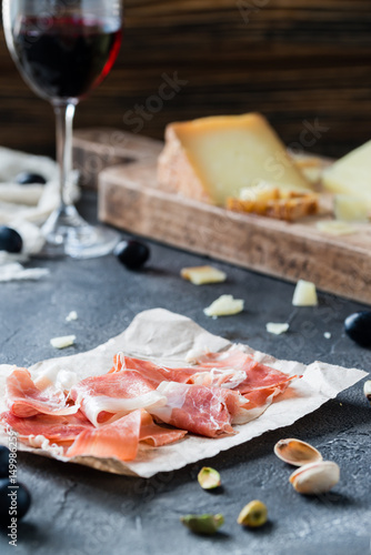 Ham jamon serrano or prosciutto crudo with sliced hard cheeses (Italian pecorino toscano and Spanish manchego), glass of red wine with grapes, pistachios, on dark rustic background