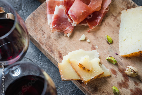 Platter with Spanish ham jamon serrano or Italian prosciutto crudo, sliced Italian hard cheese pecorino toscano, glasses of red wine and pistachios, on old wooden board