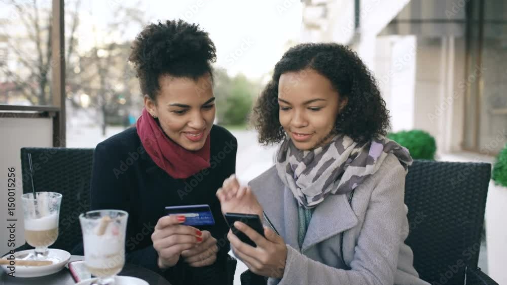 Two attractive mixed race women having online shopping with credit card and smartphone while talking and drinking coffee in street cafe Two attractive mixed race women having online shopping with credit card and smartphone while talking and drinking coffee in street cafe