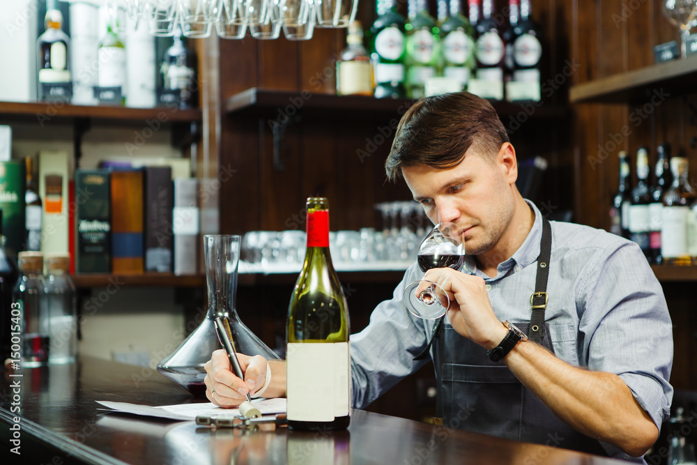Male sommelier tasting red wine and making notes at bar counter