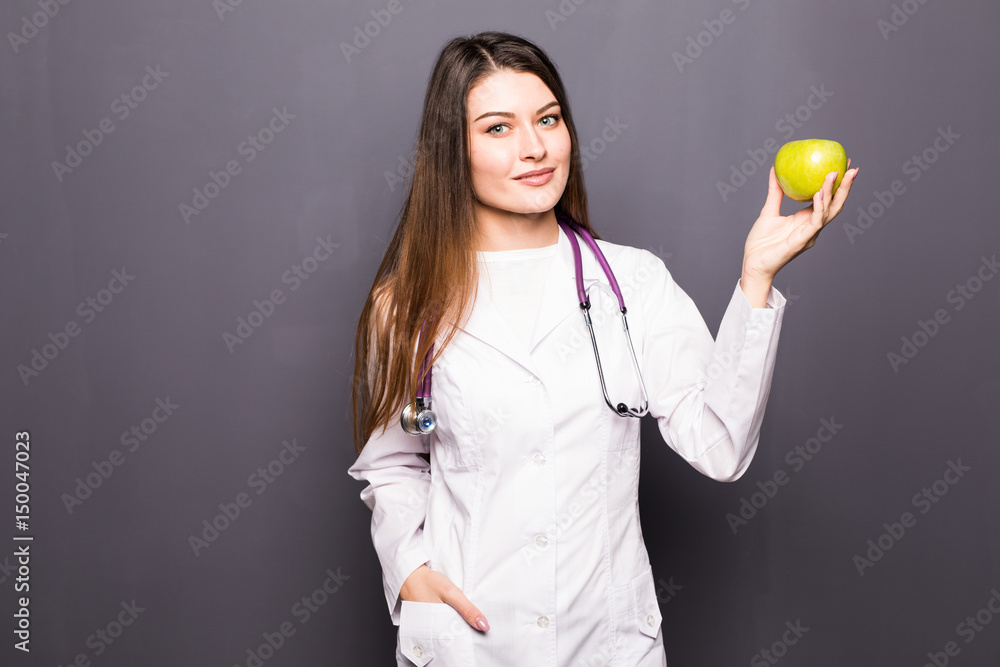 Cheerful cute young woman doctor standing and holding an apple over grey background Cheerful cute young woman doctor standing and holding an apple over grey background