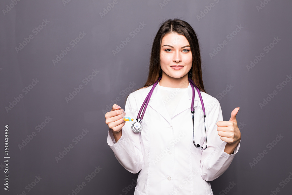 Beautiful young woman dressed as doctor holding container with pills and showing thumb up isolated over grey background Beautiful young woman dressed as doctor holding container with pills and showing thumb up isolated over grey background