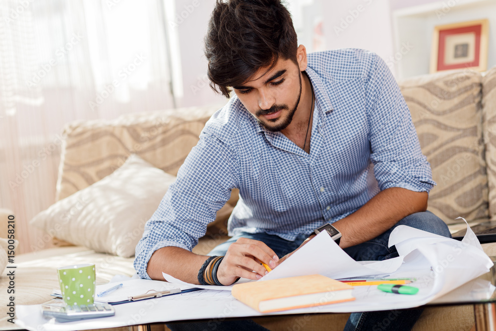Young businessman working at home. He sitting on sofa with laptop and looking at blueprints. Young businessman working at home. He sitting on sofa with laptop and looking at blueprints.