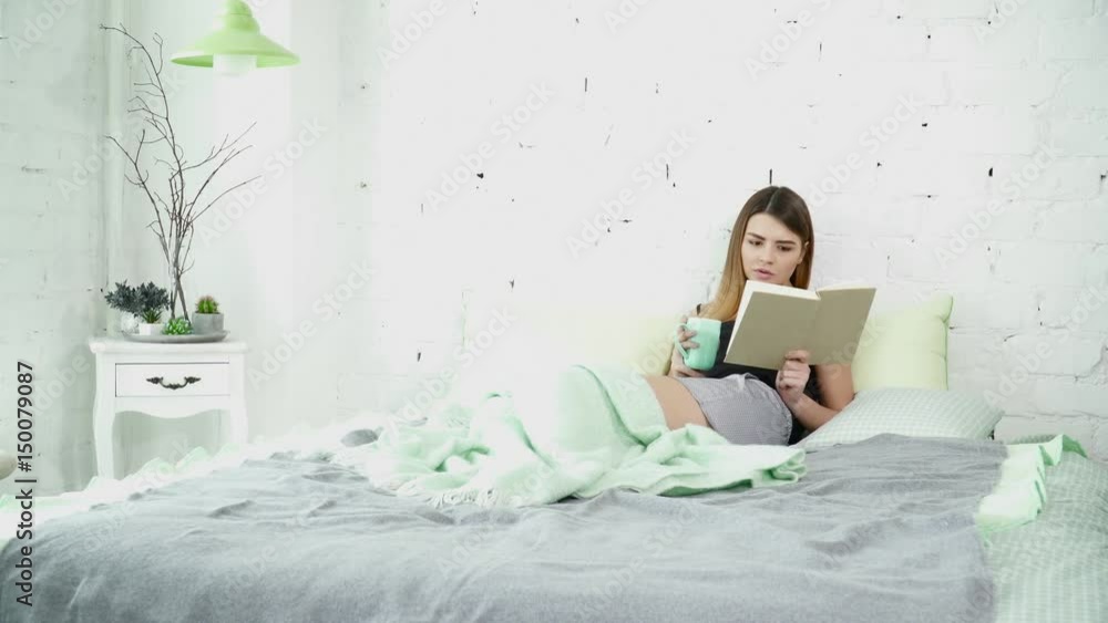 Beautiful brunette woman sitting on the bed and reading a book with a cup Beautiful brunette woman sitting on the bed and reading a book with a cup