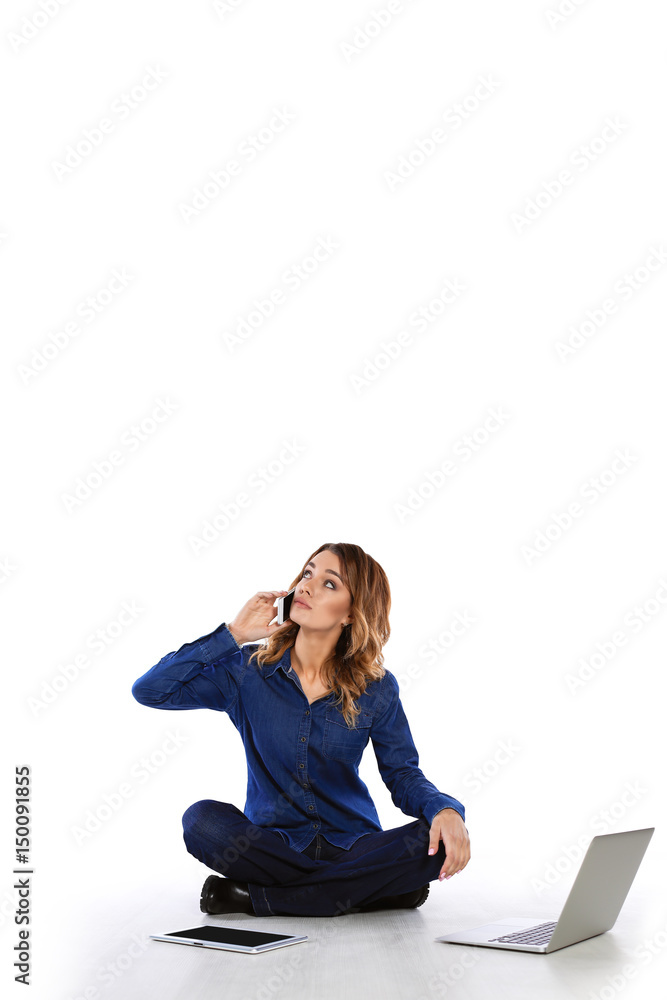 Confused woman talking on the phone while sitting on the floor with a tablet and a laptop on a white background Confused woman talking on the phone while sitting on the floor with a tablet and a laptop on a white background