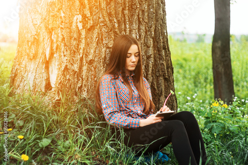 A young, pretty girl in a shirt sits near a tree on the grass on a sunny day with a notebook.