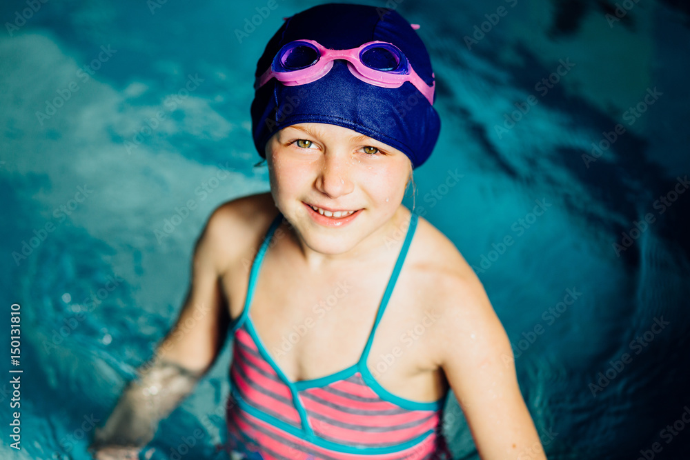 Happy little girl in blue cap and pink goggles looking up in swim pool