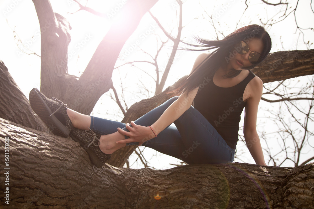 Fototapeta premium pretty young brunette woman wearing jeans and black shirt with big mirror sunglasses posing near the tree in the park