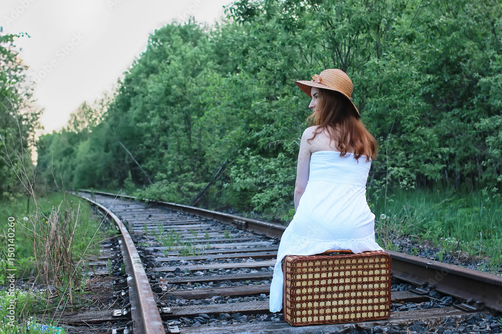 Obraz premium girl in a white sundress and wicker suitcase walking on rails