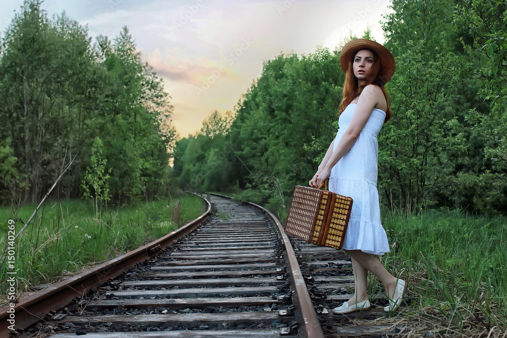 Naklejka premium girl in a white sundress and wicker suitcase walking on rails