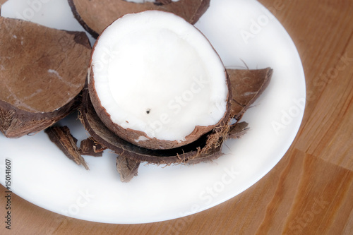 Still life with broken coconut shell ripe white flesh inside and debris on round white plate on brown wooden table horizontal close up photo