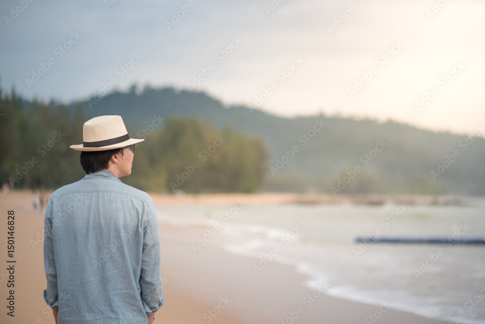 Young Asian man with jean shirt and hat walking on beautiful tropical beach and see the sunset, happy vacation time and summer holiday travel concepts