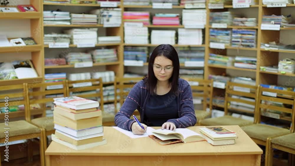 The student works through books on studies and writes notes in the library, a beautiful background of books and a lot of books on the table The student works through books on studies and writes notes in the library, a beautiful background of books and a lot of books on the table