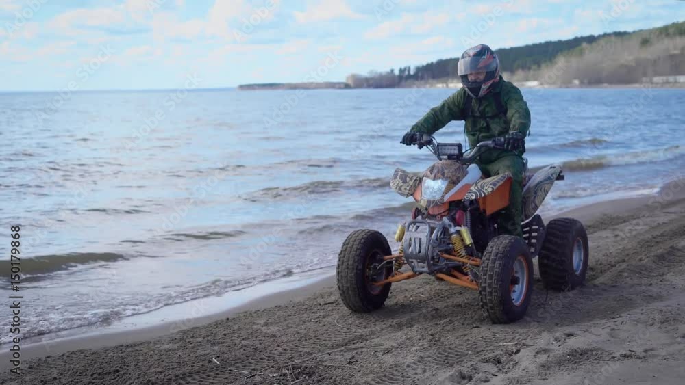 A young man is riding along the beach line near the sea on a sport quad bike with a lightweight body for a more speedy ride A young man is riding along the beach line near the sea on a sport quad bike with a lightweight body for a more speedy ride