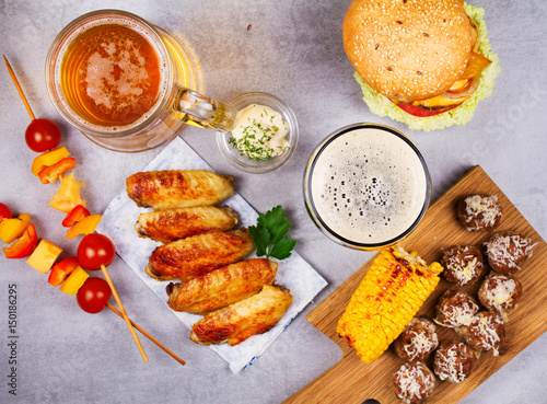 Glasses of beer with chicken wings, burger, meat balls, grilled corn and vegetables. Beer bites. Ale and food still life. View from above, top studio shot photo