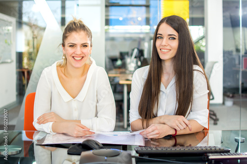 Two smiling businesswomen sitting in the office and looking at camera.