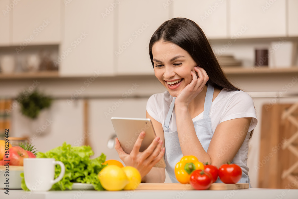 Young Woman standing in kitchen with tablet computer, looking recipes. Side view