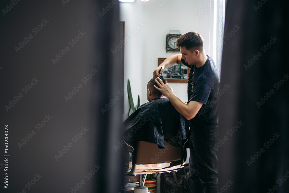 Little Boy Getting Haircut By Barber While Sitting In Chair At Barbershop. Little Boy Getting Haircut By Barber While Sitting In Chair At Barbershop.