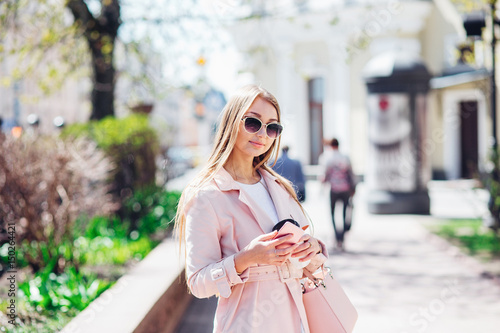 Upper class woman . Fashionable woman texting outdoors. Fashion woman in a sunglasses and pink jacket with coffee