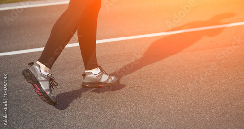 Jogging woman on track. Legs of woman running on road near park or forest. Closeup of female in running shoes going for run on road at sunrise or sunset. Toned.
