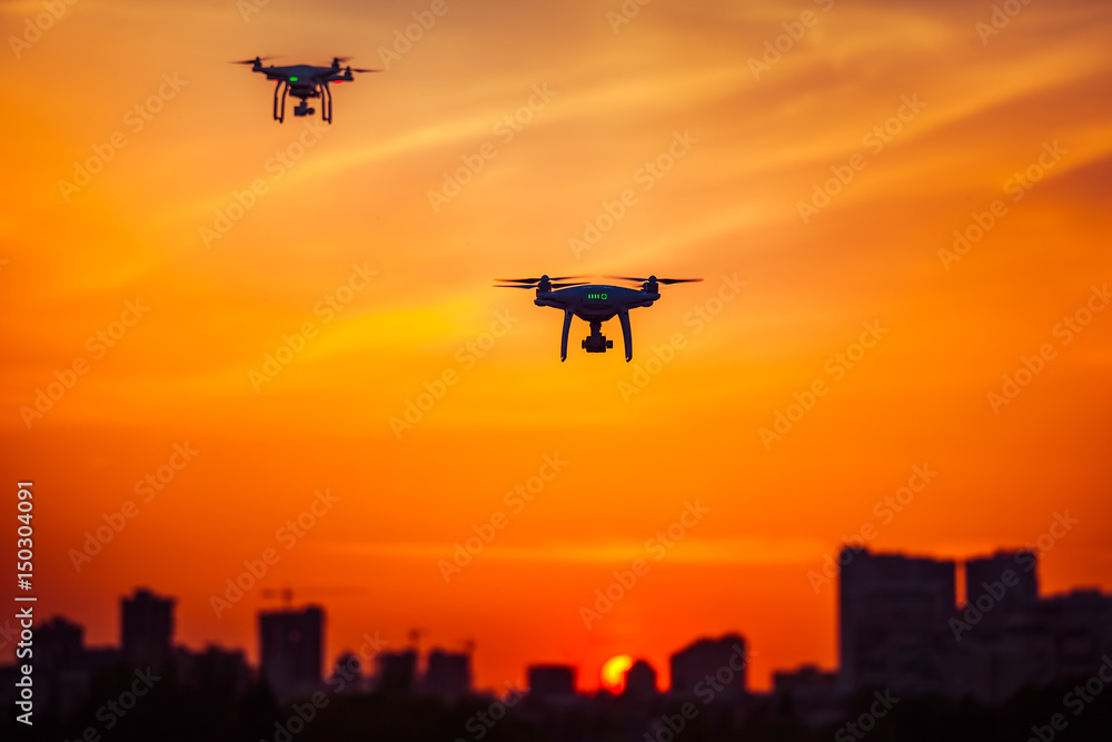 Fototapeta premium Two modern Remote Control Air Drones Fly with action cameras in dramatic orange sunset sky. Cityscape silhouette in the background. Modern technologies. Kiev, Ukraine. Travel, hobby, inspiration