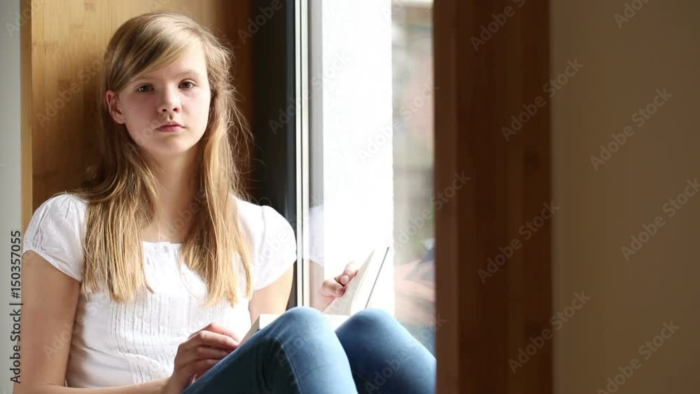 Young girl sitting on window sill reading a book, looking up Young girl sitting on window sill reading a book, looking up