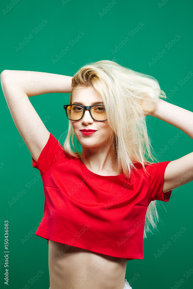 Smiling Girl with Long Blond Hair and Eyeglasses Wearing Short Red Top is Posing in Studio on Green Background. Smiling Girl with Long Blond Hair and Eyeglasses Wearing Short Red Top is Posing in Studio on Green Background.