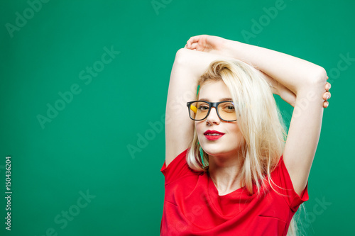 Portrait of Girl with Long Blond Hair, Eyeglasses and Charming Smile in Red Top Posing on Green Background in Studio.