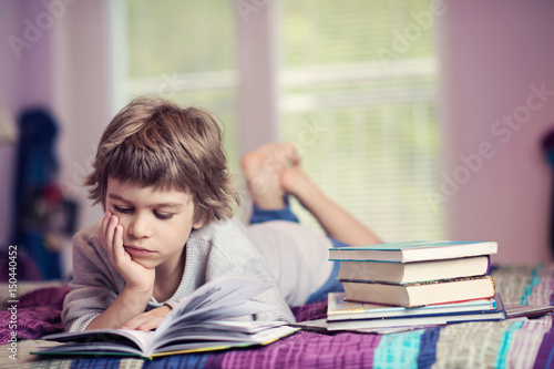 Cute little boy lying on bed reading next to stack of books