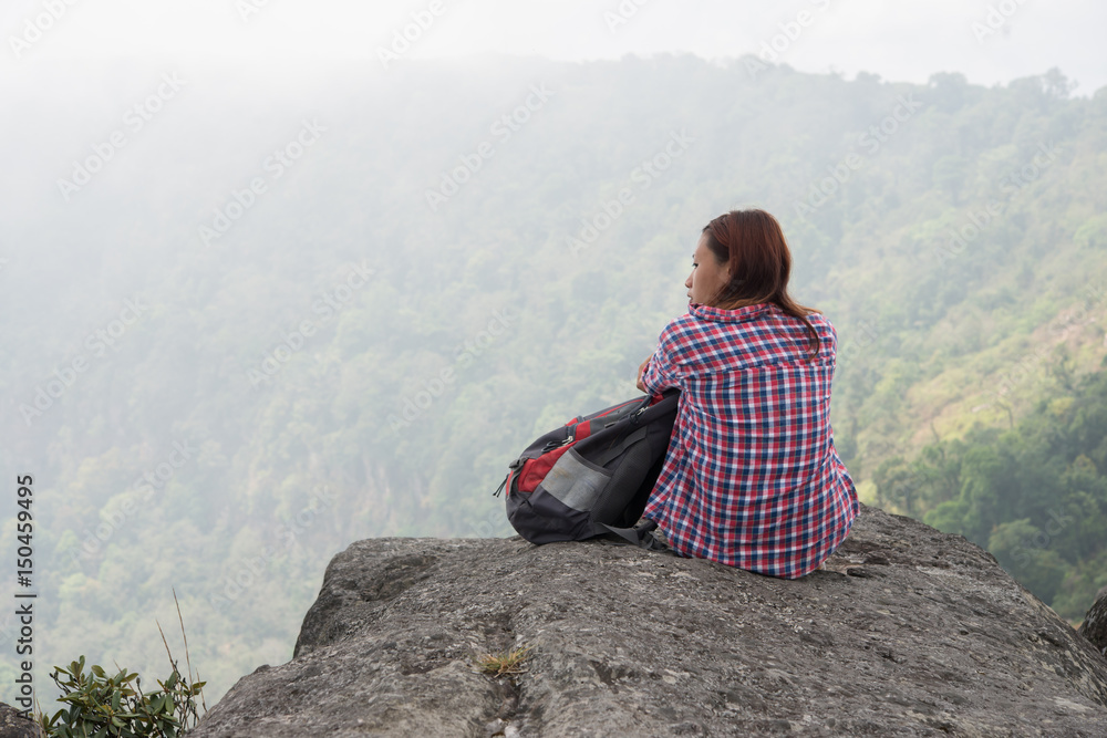 Young hiker with backpack sits on the edge of the cliff and looking at the nature valley and mountains.