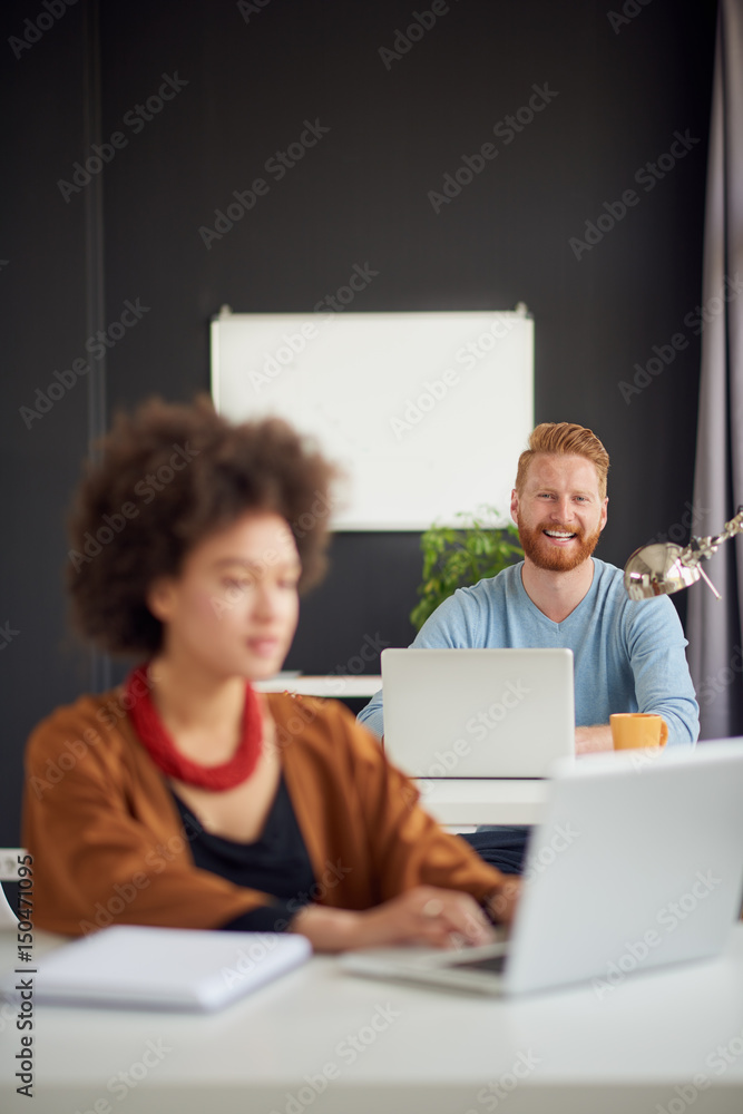 Young Caucasian businessman sitting and using laptop computer in modern office. In background colleague Young Caucasian businessman sitting and using laptop computer in modern office. In background colleague