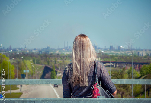 Woman with blond long hair observing cityscape – road and smoke from factories chimneys, standing on a  bridge © elenakirey