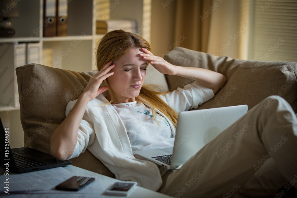 Attractive businesswoman on a couch with two cellphones and laptops. Exhausted woman in office late at night. Responsible executive working, having headache, hands on temples Attractive businesswoman on a couch with two cellphones and laptops. Exhausted woman in office late at night. Responsible executive working, having headache, hands on temples