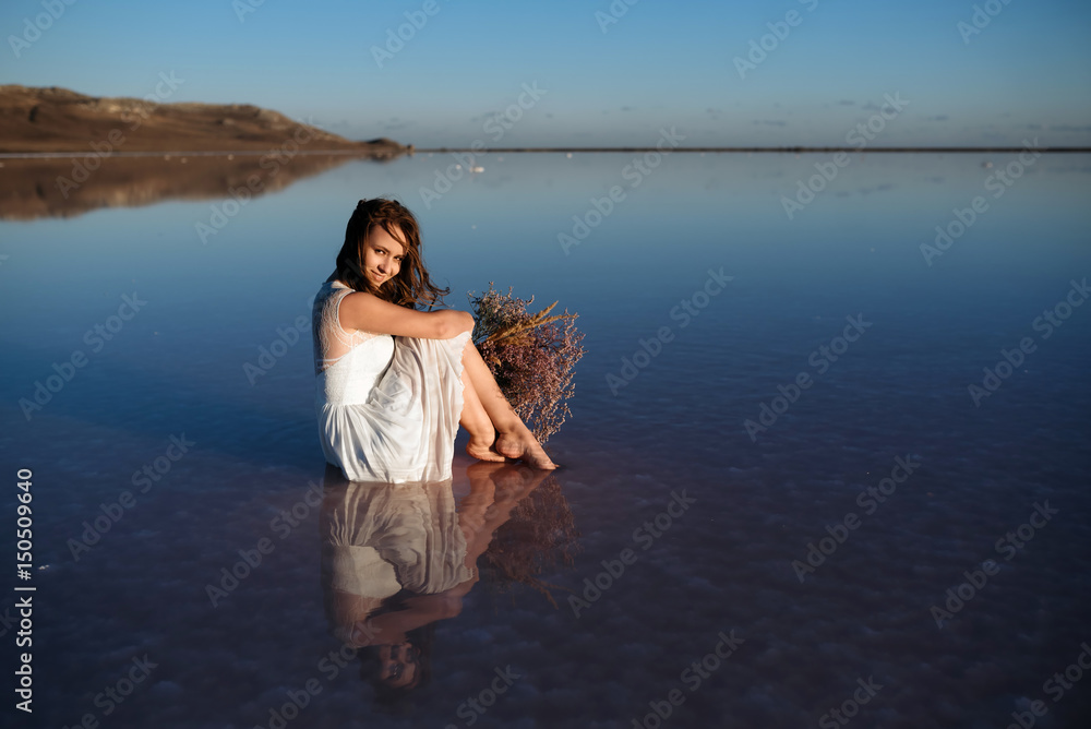 Girl in white dress sitting in salt lake with flowers at sunset. look like bride