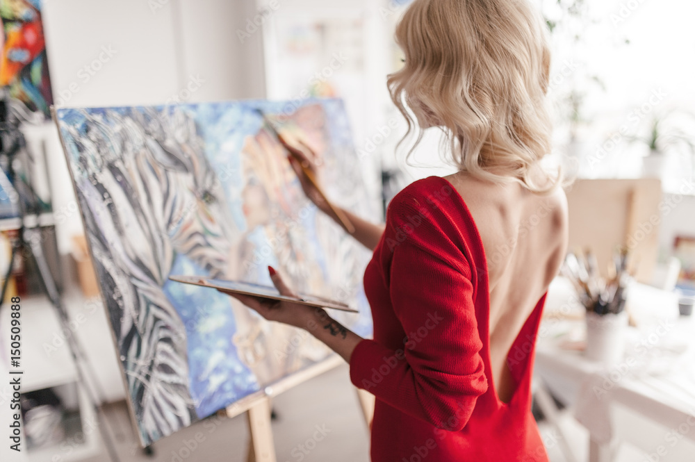 The artist in a red dress stands and paints with a brush on canvas.