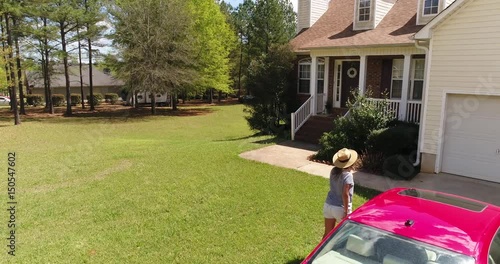 Drone video 4K 60 fps. Young woman opens door, gets out of the red car and comes  inside the house. Girl returning home from work or shopping. Beautiful suburb house located in green subdivision
