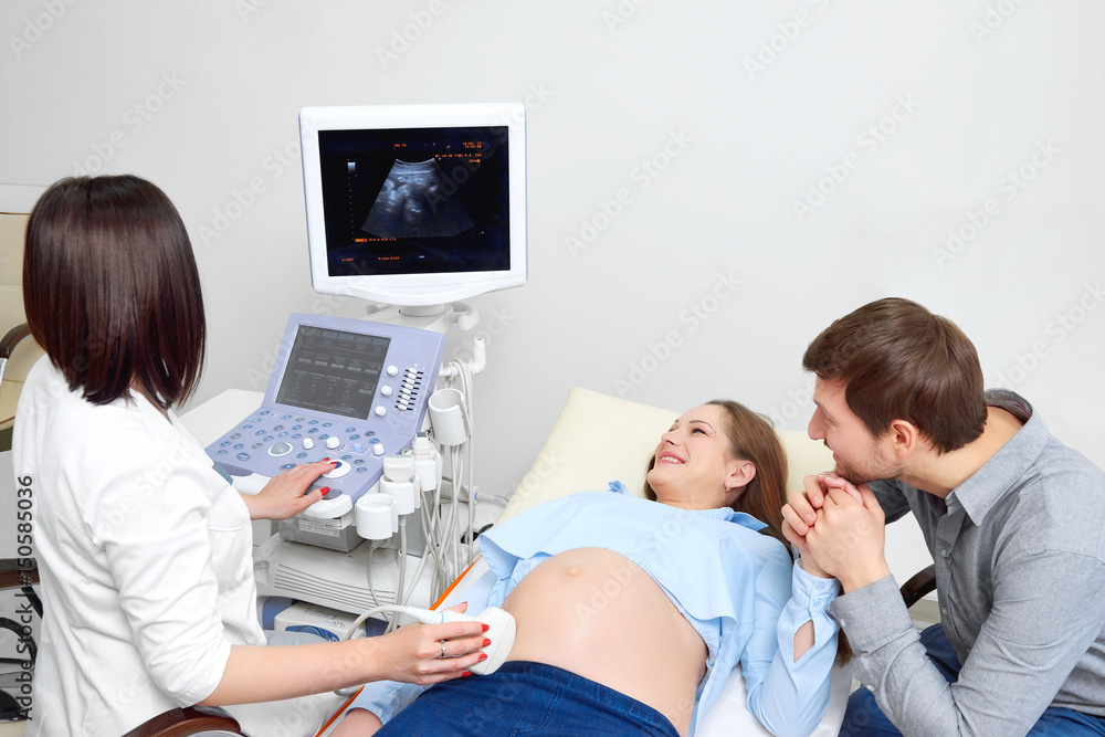 Young loving pregnant couple looking excited while visiting doctor together looking at the monitor with a sonogram of their unborn child medicine healthcare people professional experience qualified. Young loving pregnant couple looking excited while visiting doctor together looking at the monitor with a sonogram of their unborn child medicine healthcare people professional experience qualified.