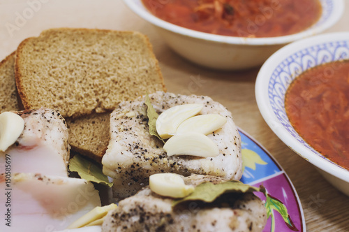 Salted lard with pepper and bay leaf and black bread lies on a plate, next to it there are two plates with red borsch.. Close-up, selective focus.