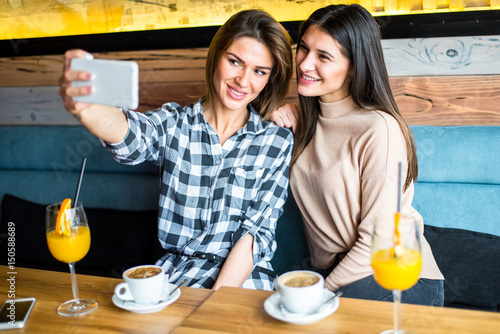 Two pretty young women sitting by the table and taking selfie in cafe