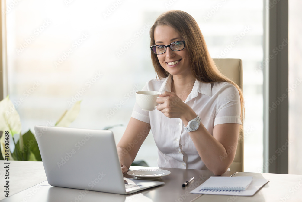 Businesswoman relaxing with cup of aromatic coffee. Smiling confident girl sitting at the desk with mug of hot tea in hand. Short break at the middle of hard working day concept. Businesswoman relaxing with cup of aromatic coffee. Smiling confident girl sitting at the desk with mug of hot tea in hand. Short break at the middle of hard working day concept.